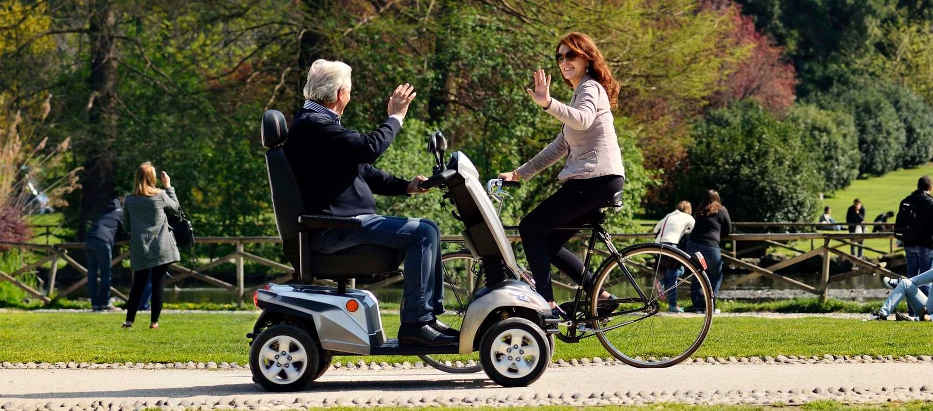 Ein Mann und eine Frau fahren auf einem Roller in einem Park. Bäume und Wiesen sind im Hintergrund sichtbar.